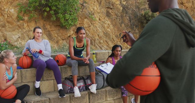 Female basketball team receiving coaching outdoors. Women in athletic wear holding basketballs, listening to coach with clipboard. Perfect for sports training, teamwork, motivational materials, and outdoor fitness concepts.