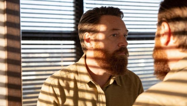 Bearded man studying reflection through blinds in tan shirt with dramatic striped sunlight