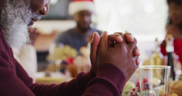 Elderly man praying at a festive Christmas dinner table with family members in the background. Hands are folded in prayer, expressing gratitude and enjoying the holiday celebration. Ideal for use in content related to family gatherings, holiday traditions, Christmas celebrations, spirituality, and expressions of gratitude.