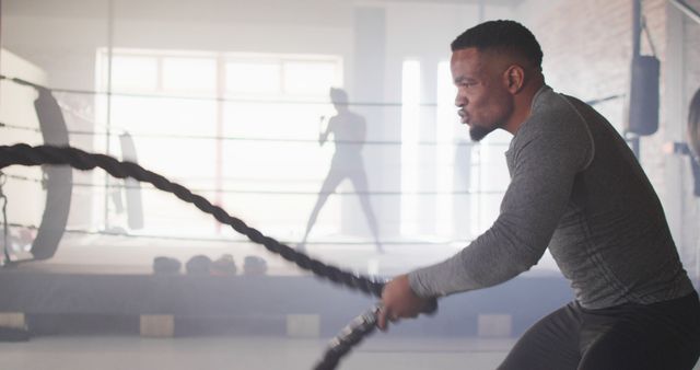 A man is using battle ropes during his workout session in an indoor gym. He appears dedicated and focused. This image is ideal for illustrating fitness training, promoting athletic gear, or motivational content related to exercise and physical conditioning.