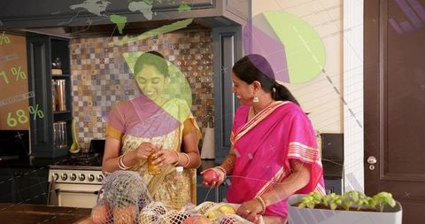 Two indian women preparing pomegranate at kitchen island wearing gold and pink sarees