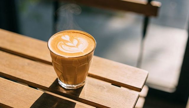 Steaming glass latte with heart foam art on sunlit wooden table morning cafe calm