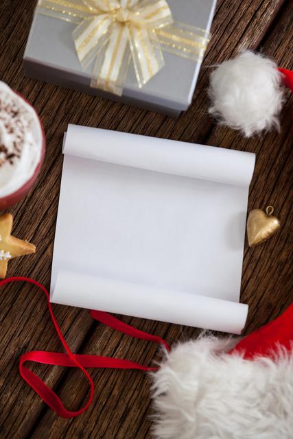 Blank white paper positioned with a festive Santa hat, golden gift, and red ribbon on a rustic wooden table. Ideal for use in holiday-themed projects, Christmas card designs, or promotional materials for festive events. Perfect for conveying a cozy, warm holiday atmosphere and planning for the Christmas season.