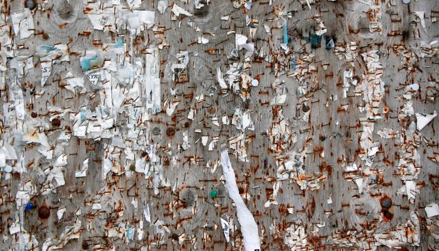 This image depicts an old bulletin board showing the remnants of torn posters and rusty nails. Rust and tattered paper fragments cover the rough surface of the board, creating a textured look. It illustrates themes of urban decay, neglect, and vintage aesthetic. This image is ideal for use in artworks, urban decay themed projects, backgrounds, or editorial pieces exploring city life and deterioration.