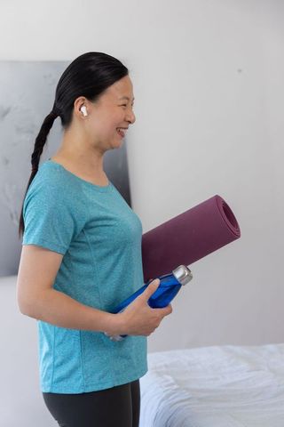 Mature asian woman smiling holding yoga mat and water bottle wearing wireless earbuds