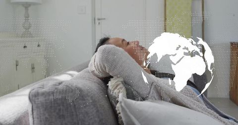 Relaxing asian man reclining on sofa with global map overlay in minimalist living room