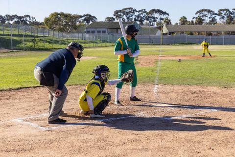 Youth baseball batter swinging at incoming pitch with catcher and umpire on sunny field