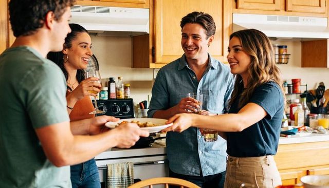 Young friends passing plate and sharing drinks in cozy home kitchen during casual dinner