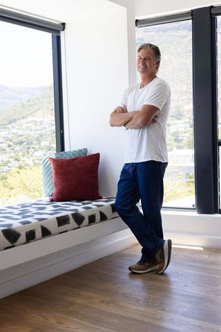 Mature man relaxing in sunlit modern window nook with bench and pillows, hillside view