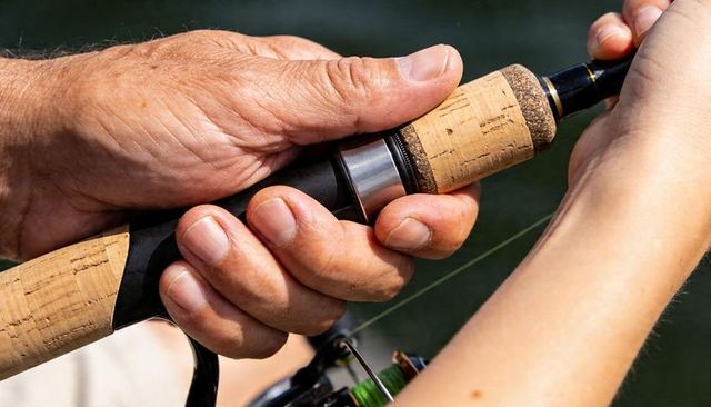 Senior hand guiding child holding cork-handled spinning rod closeup at lake, mentoring moment