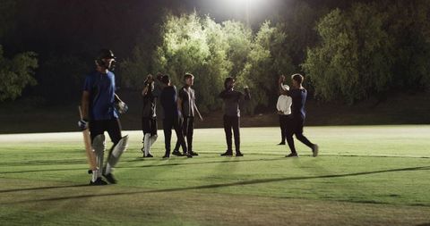 Night cricket team practicing under floodlights, batsman walking off in pads and helmet