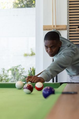 Young african american man leaning over pool table aiming cue in sunlit home game room