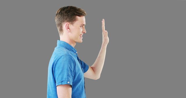 Man in casual blue shirt gesturing with hand, indicating hello or goodbye. Neutral gray background emphasizes the subject, making this useful for presentations, communication advertisements, and social media campaigns promoting friendliness and approachability.