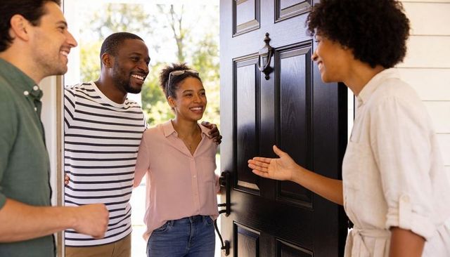 Homeowner welcoming smiling guests at front door with open black door and porch
