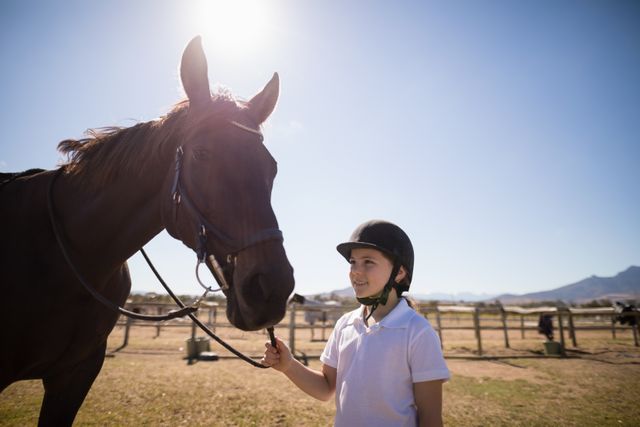 This image depicts a smiling girl standing next to a horse, holding its rein, while wearing a riding helmet. The setting is an outdoor ranch on a sunny day, with mountains visible in the background. Ideal for use in content about equestrian activities, children's outdoor hobbies, ranch life, animal bonding, or promoting horse riding education.