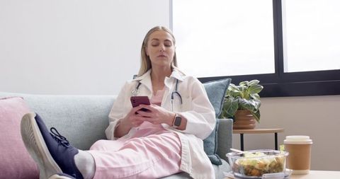 Nurse relaxing on sofa in clinic lounge holding smartphone and scrolling