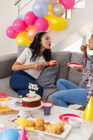 Diverse young women celebrating 18th birthday on couch with balloon arch and cake