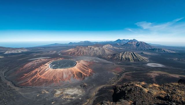 Reddish cinder cone crater rising from black basalt plain with distant volcanic range
