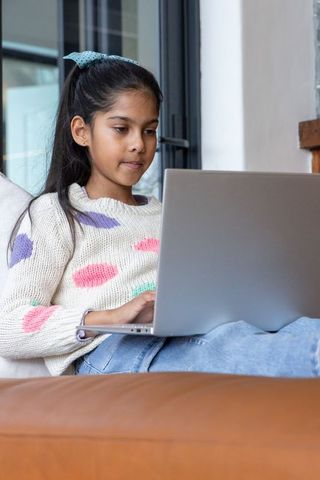 Indian girl using laptop on sofa studying online and learning at home, remote education focus
