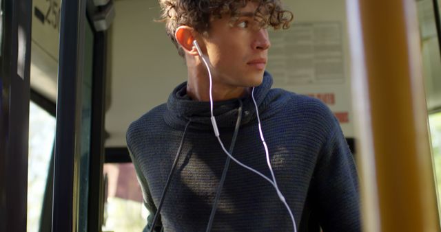 Young man with curly hair wearing a sweater and headphones, standing on public transport during the day and looking out of the window with a thoughtful expression. Ideal for use in content related to modern commuter life, urban transport, technology, and youth culture.