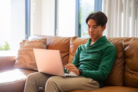 Young asian man working on laptop on tan leather couch in bright modern living room