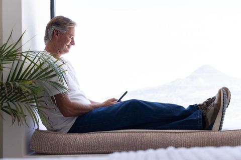 Mature man reclining on cushioned window seat using smartphone, sunlit interior and mountain view