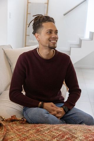 Smiling man sitting cross-legged on sofa wearing burgundy sweater in modern living room