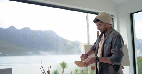 Indian artist mixing paint in wooden bowl by coastal studio window with mountain view