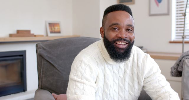 A man is smiling while relaxing at home on a comfortable chair. He is wearing a cozy sweater and appears content in his living room setting. This image could be used for lifestyle blogs, articles on home comfort, advertisements for home furnishings, or any content promoting a relaxed and happy lifestyle.