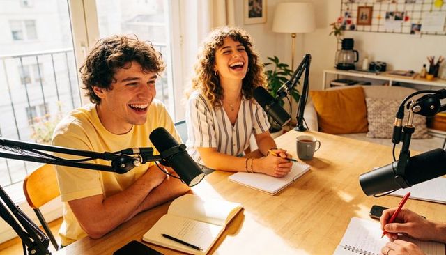 Young co-hosts laughing while recording podcast at sunlit wooden table with pro mics