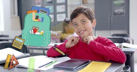 Young boy raising hands in classroom using tablet surrounded by colorful school supplies