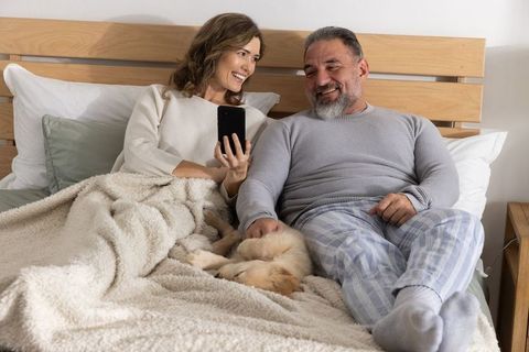 Middle-aged couple relaxing in bed with puppy, woman showing smartphone, cozy morning