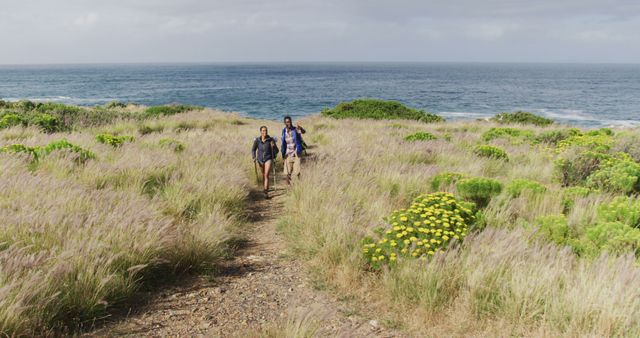 Couple hiking on scenic coastal trail surrounded by lush greenery and wildflowers, with ocean view in background. Perfect for promoting outdoor adventures, travel destinations, nature activities, and healthy living.