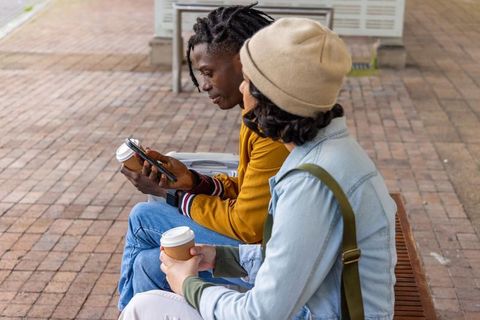 Young commuters checking smartphone, holding coffee while waiting on transit platform with suitcase