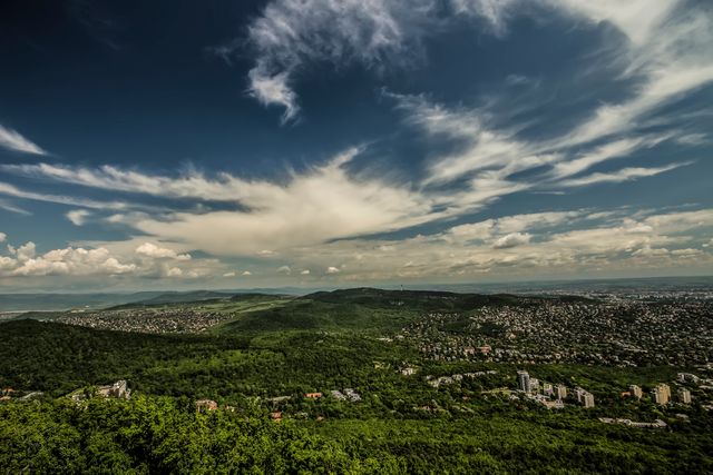 Expansive view of lush green hills contrasting with a distant cityscape under a dramatic sky with clouds. This image can be used for backgrounds, travel promotions, nature and city editorial content, and environmental awareness campaigns.