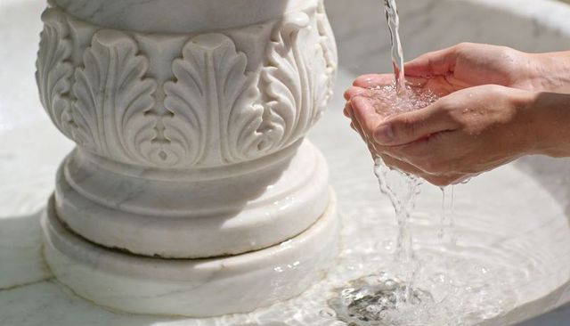 Cupped hands catching flowing water from marble fountain basin in sunlit plaza