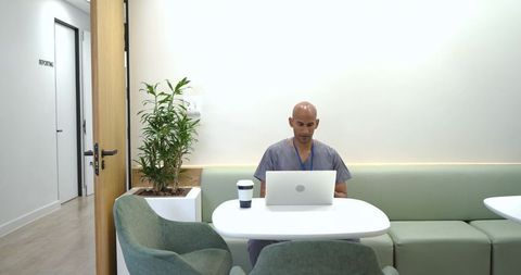 African american healthcare worker in scrubs working on laptop in modern clinic breakroom