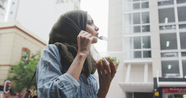Thoughtful biracial woman in hijab walking in city street eating takeaway salad. City living and healthy modern urban lifestyle.