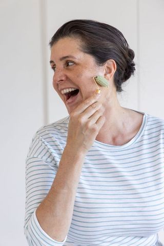 Mature woman using green jade facial roller, smiling in at-home skincare self-care routine