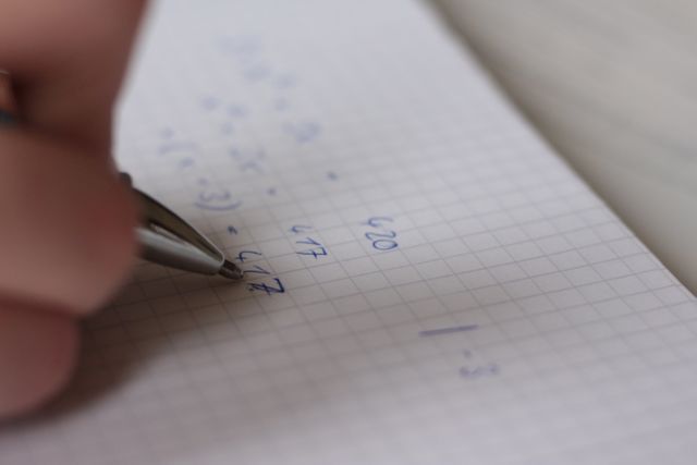 Photo showing a close-up of a hand holding a pen and solving a mathematics problem in a notebook. Suitable for educational content, math tutorials, student study guides, and articles on learning or homeschooling techniques.
