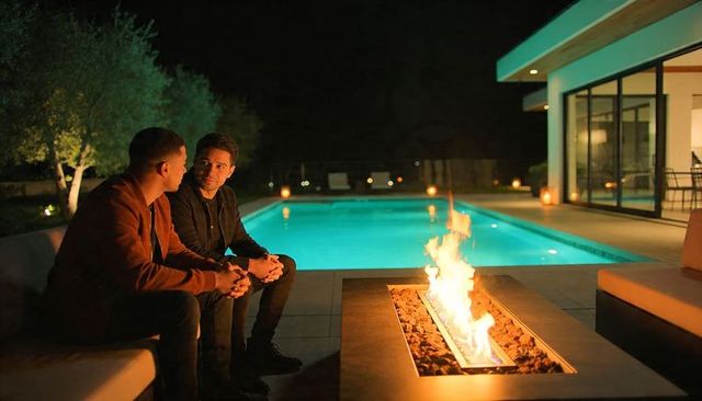 Two men talking by modern pool and fire pit at night, intimate outdoor patio ambience