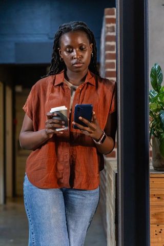 Young professional leaning on office counter checking smartphone holding takeaway coffee