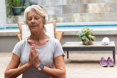 Senior woman meditating on poolside patio wearing headphones and smartwatch for calm