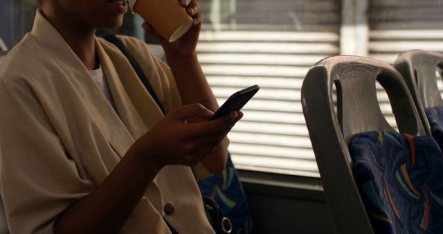 African american woman sitting in city bus drinking takeaway coffee and using smartphone. Communication, transport, city living and lifestyle, unaltered.