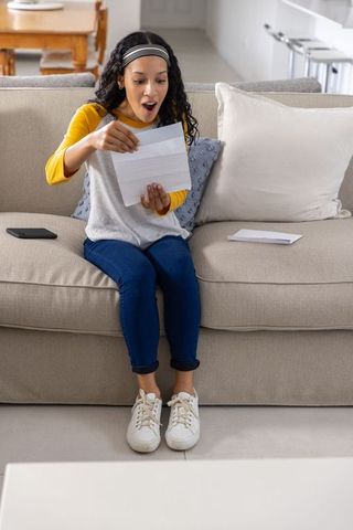 Surprised teen girl opening letter on couch, reacting with joyful shock at home
