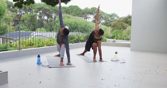 Two men are practicing yoga on a spacious terrace overlooking a lush green landscape. They are dressed in athletic wear and are performing a stretching pose on yoga mats, with water bottles nearby hinting at their commitment to staying hydrated. This image can be used in promotional content for fitness retreats, yoga classes, wellness programs, or outdoor exercise activities.