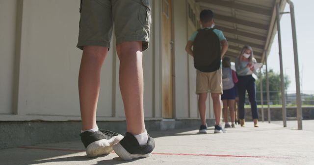 Students are standing in a line outside a schoolwhile maintaining proper social distancing by adhering to the marked lines. They are wearing masks, a measure for preventing the spread of COVID-19. This image can be used in articles and blogs related to education during the pandemic, safety measures, school reopening, and child health.