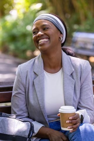 Smiling african woman in gray blazer laughing and holding takeaway coffee on park bench