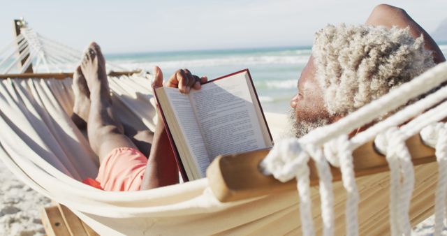 Senior african american man reading and lying in hammock on sunny beach. healthy and active time beach holiday.
