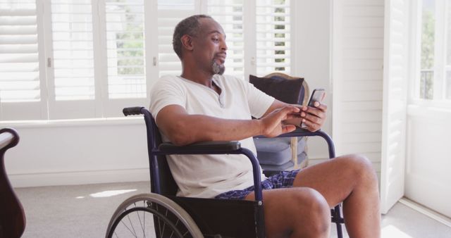 Smiling senior african american man sitting in wheelchair at home using smartphone. Communication, senior lifestyle, disability and domestic life.